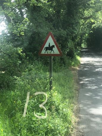 A photograph of a triangular horse riding warning sign on the side of a rural lane with a white number 13 painted on the grass.
