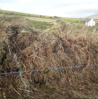 A photograph showing a grassy field with a wire fence in the foreground and a rural landscape with stone walls and a white house in the background.