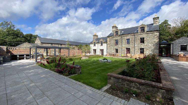 A wide-angle photograph showing a large stone detached house with a landscaped rear garden, paved patio, and pergola structure under a blue sky.
