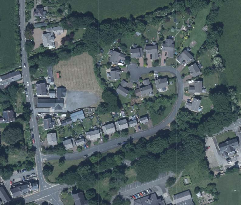 An aerial satellite view showing a rural residential area with a road network, existing houses, and a large undeveloped plot of land.