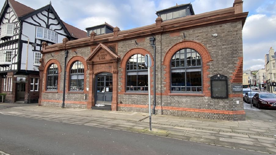 A street-level photograph of the Old Market Hall, a historic grey brick building with red brick arches and large windows, located next to a timber-framed structure.