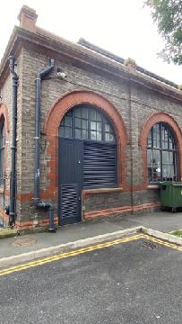 A photograph showing the exterior brick facade of a building with large arched openings, one containing a black door and another a window.