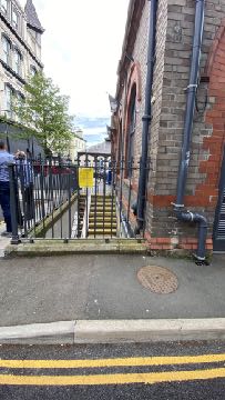 A street-level photograph showing the exterior of a brick building with a black metal fence and gate in the foreground. Several people are standing near the fence on the left side of the frame.