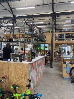Interior photograph of a commercial space, likely a shop or cafe, featuring wooden shelving, a counter, and bicycles in the foreground.
