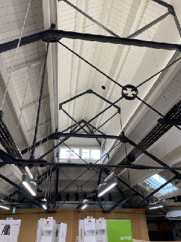 The image shows an interior perspective looking up at a complex black metal truss framework suspended from a white ceiling with skylights. Display boards are visible at the bottom, suggesting a commercial or public in...