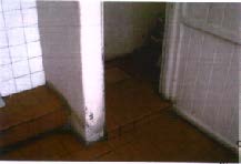 A low-resolution interior photograph showing a corner with white square tiled walls, a wooden step or floor section, and a white door or paneling.