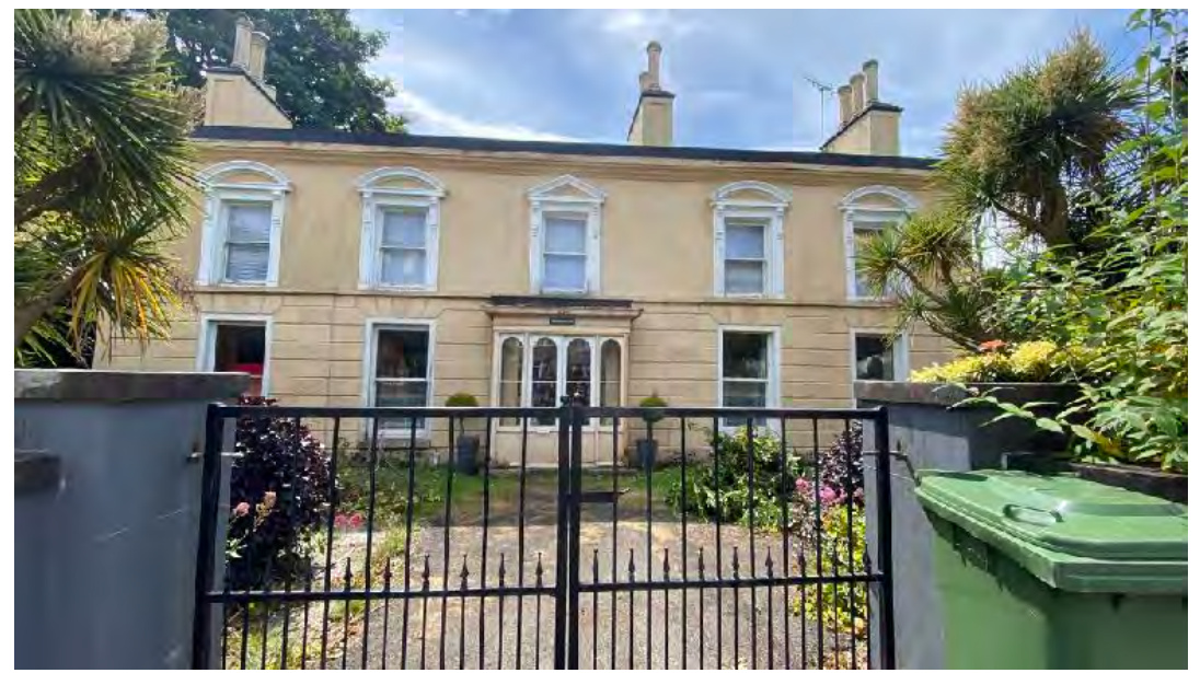 A street-level photograph of a large, two-story cream-colored terraced house with white window frames and a black metal gate in the foreground.