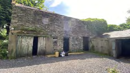 A photograph showing a large, weathered stone building with multiple wooden doors, resembling a barn or agricultural outbuilding on a gravel site.