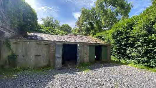 A photograph showing a small, detached outbuilding or garage with a tiled roof and green doors, located on a gravel driveway surrounded by dense vegetation.
