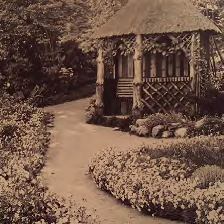 A sepia-toned photograph showing a rustic thatched garden structure or summer house with wooden lattice walls, situated alongside a paved path in a garden.