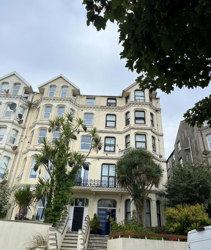 A low-angle photograph of a large, cream-colored, multi-story building with bay windows and balconies, partially obscured by palm trees in the foreground.