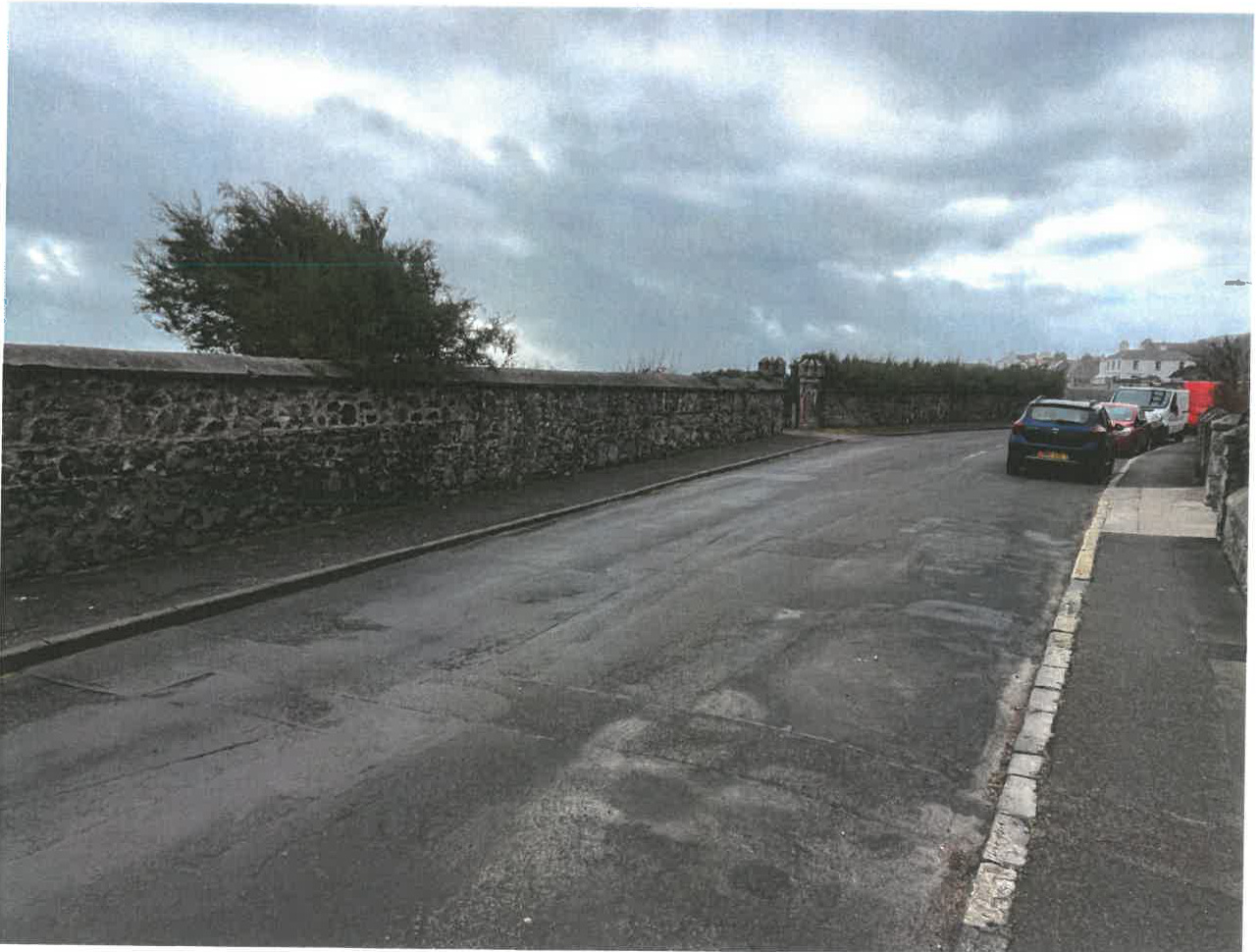 A street-level photograph showing a paved road bordered by a long stone wall on the left and parked cars on the right, likely depicting the existing access point.