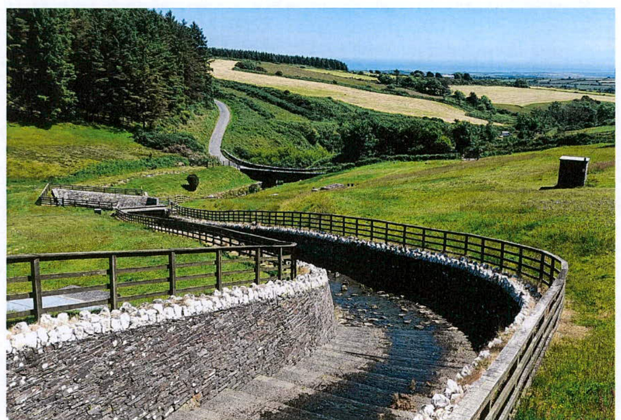 A photograph showing a stone spillway channel with wooden fencing running through a grassy, hilly landscape with fields and trees in the background.