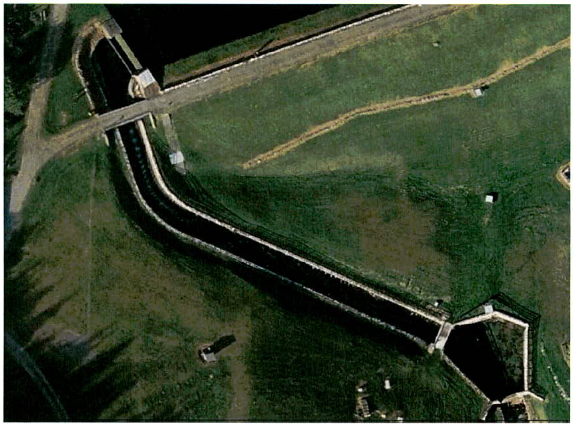An aerial photograph showing a long concrete spillway channel running through a grassy field, consistent with the Cringle Impounding Reservoir.