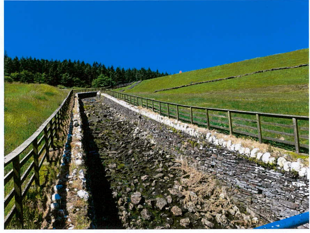 A photograph showing a stone-lined spillway channel running down a grassy hillside with wooden railings on either side.
