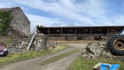 A photograph of a rural site featuring an old stone building on the left and a large open-sided agricultural outbuilding in the background, with a dirt track in the foreground.