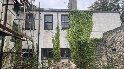 A photograph of a two-story white building with black windows, partially covered in ivy, with scaffolding on the left and a pile of rubble at the base.