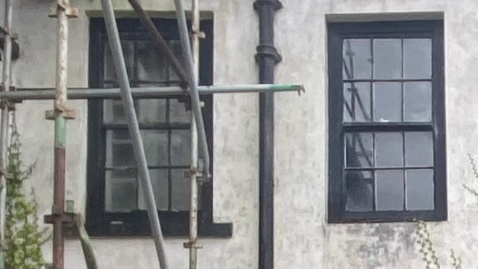 A close-up photograph of a building's exterior wall featuring two black-framed sash windows and a vertical drainpipe, with metal scaffolding visible on the left side.