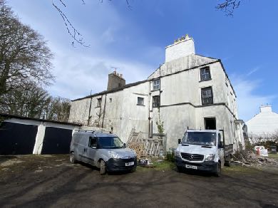 A photograph showing a large, white-washed, multi-story farmhouse with a chimney, situated in a rural setting with vehicles parked in the foreground.