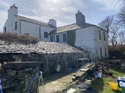 A photograph of a large white farmhouse with slate roofs and chimneys, featuring a lower stone outbuilding in the foreground.