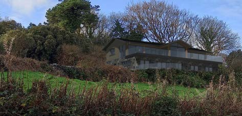 A photograph showing a large detached house situated on a grassy slope surrounded by trees and vegetation.
