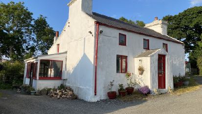 A colour photograph showing the exterior of a white, two-storey detached farmhouse with red window frames and a gravel driveway.