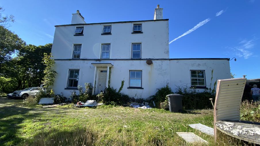 A sunny exterior photograph of a white two-story detached house featuring a single-story extension on the right side, with a grassy garden and parked cars in the foreground.