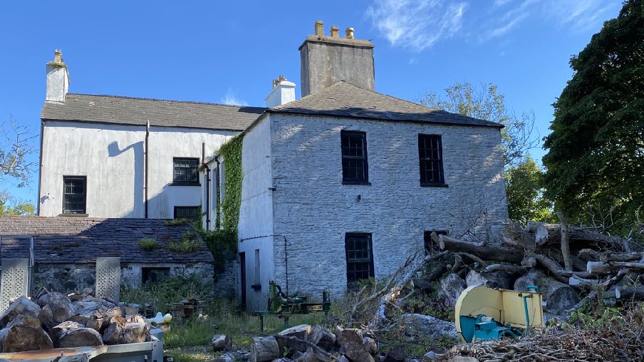 A photograph of a large, white-washed stone dwelling with slate roofs and chimneys, situated in a rural setting with timber piles in the foreground.
