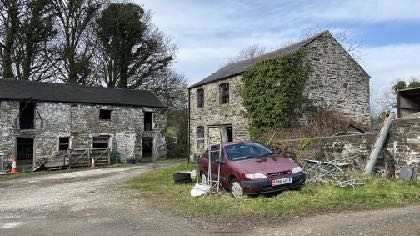 A photograph of a rural property featuring stone buildings, including a two-story house and a single-story outbuilding, with a red car parked in the foreground.