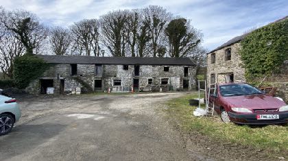 A photograph showing a long stone building, likely a converted barn, with a separate stone structure to the right. There is a gravel driveway with parked cars in the foreground.