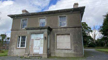 A photograph showing a two-story, weathered building with a white door and boarded window, situated in a rural setting.
