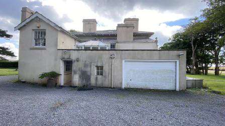 A photograph of an existing single-story building featuring a large white garage door and a two-story section, set on a gravel driveway in a rural environment.