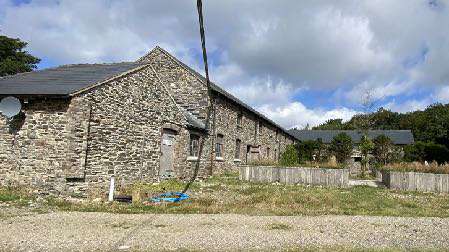 A photograph showing a large, traditional stone barn or agricultural building situated on a rural site with gravel and grass in the foreground.