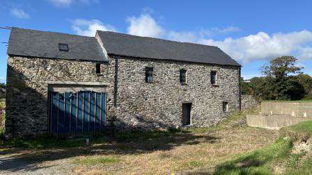 A photograph of a large, traditional stone agricultural building with a slate roof and a modern blue gate, situated in a rural grassy field.