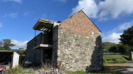 A photograph showing a dilapidated stone barn or outbuilding with a damaged roof structure, situated in a rural setting with grass and trees in the background.