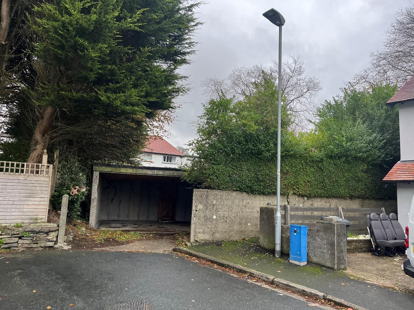 A street-level photograph showing an existing concrete garage structure next to a wooden fence and a tall street lamp post.