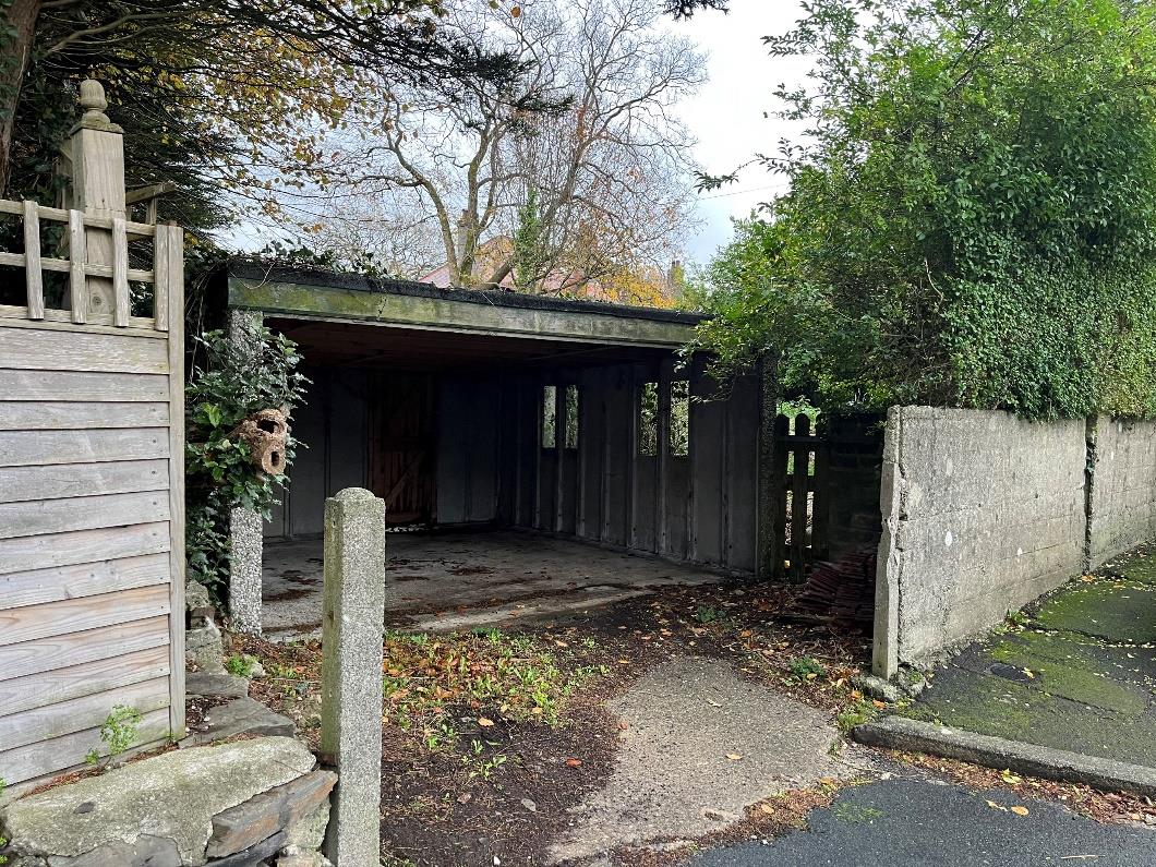 A photograph showing an existing concrete garage or carport structure with a flat roof, situated between a wooden fence and a concrete wall.