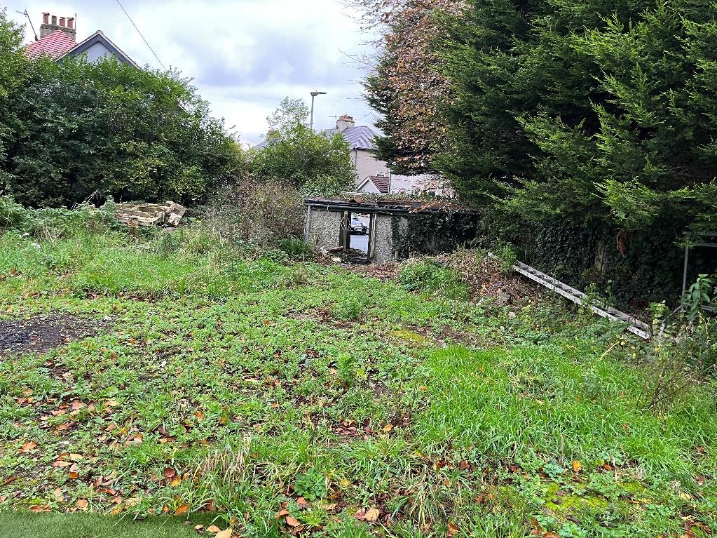 A photograph of an overgrown grassy plot of land featuring a small, dilapidated shed or outbuilding in the center, with residential houses visible in the background.