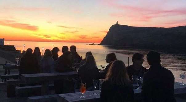 A photograph showing a group of people seated at outdoor tables on a terrace overlooking the sea at sunset, with a prominent cliff in the background.