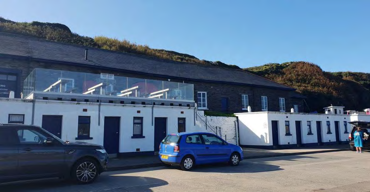 A street-level photograph showing a white terraced building with a glass-enclosed deck on the upper level, with parked cars in the foreground and a grassy hill behind.