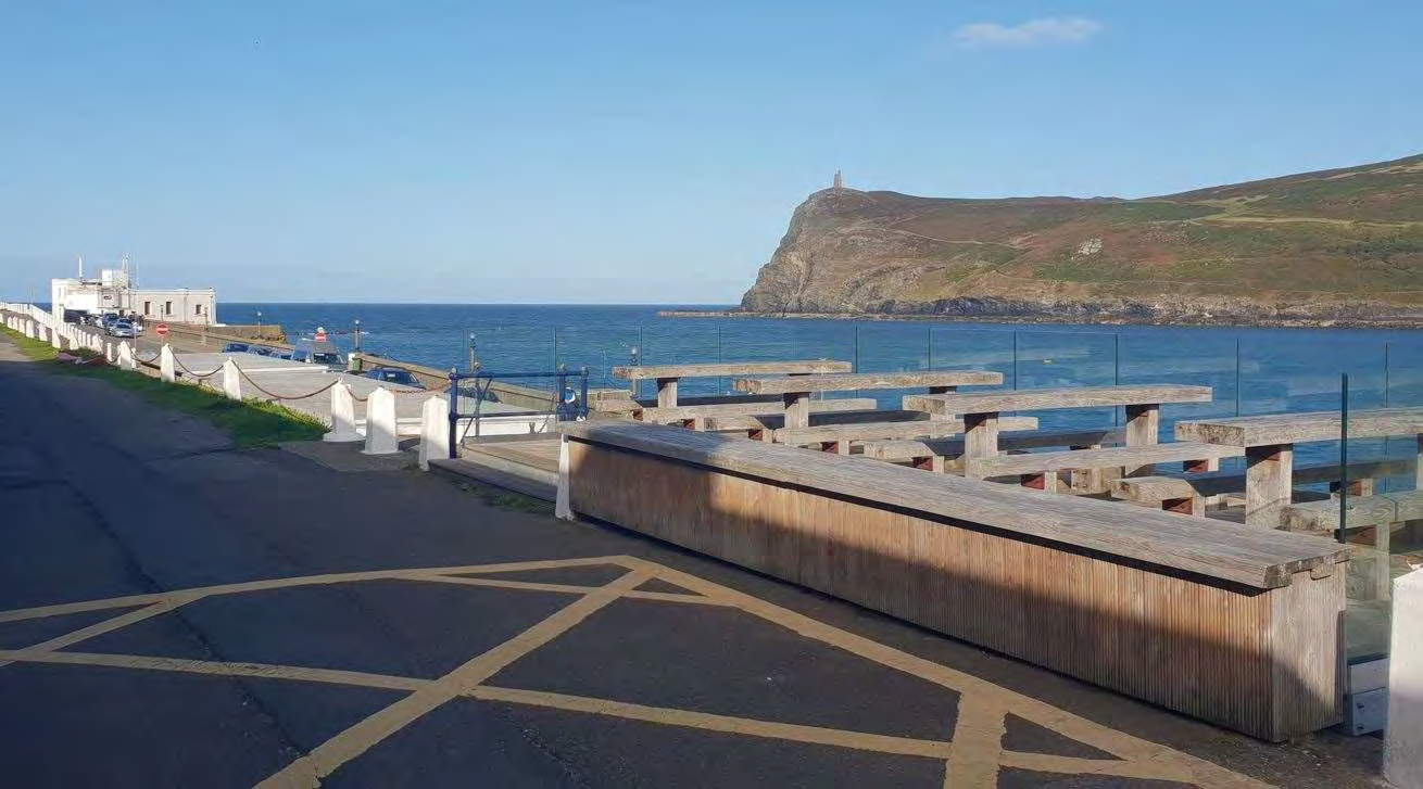 A photograph of a coastal promenade featuring wooden picnic tables and benches overlooking the sea and a headland.