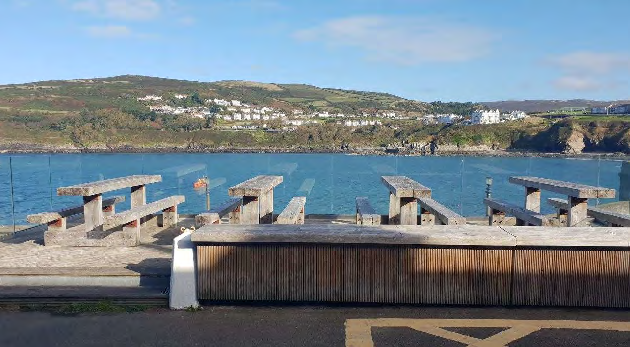 A photograph showing a wooden deck with picnic tables and glass railings overlooking a bay with a coastal village in the background.