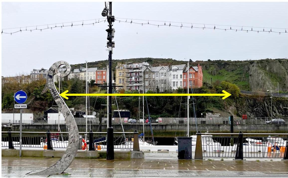 A photograph of a waterfront harbor area featuring a large metal sculpture in the foreground and a row of colorful buildings on a hillside in the background. A yellow double-headed arrow is overlaid across the middle...