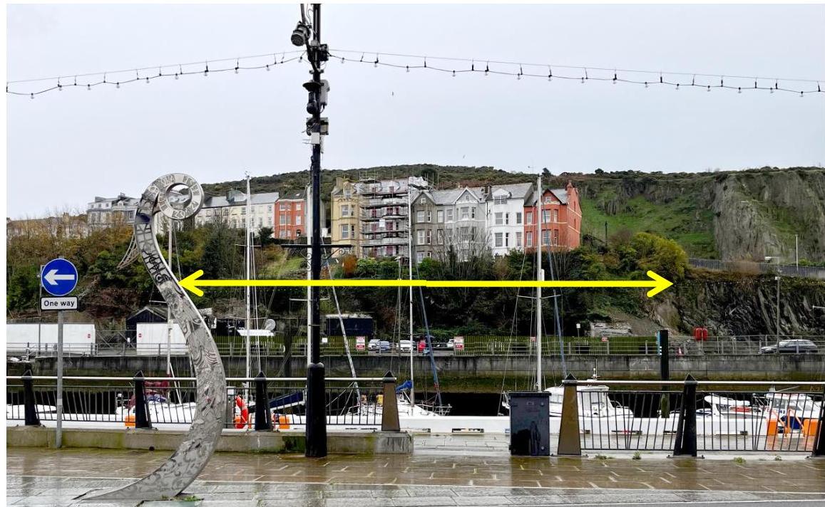 A photograph of a waterfront area in Douglas showing a harbor with boats and buildings on a hillside. A yellow double-headed arrow spans across the middle ground, indicating the extent of the site.