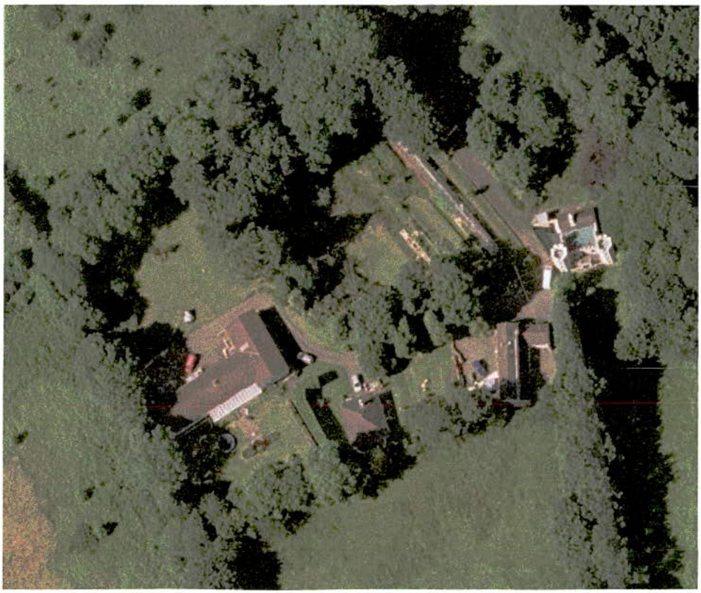 An aerial view of a rural property showing a main house, outbuildings, and dense surrounding vegetation.