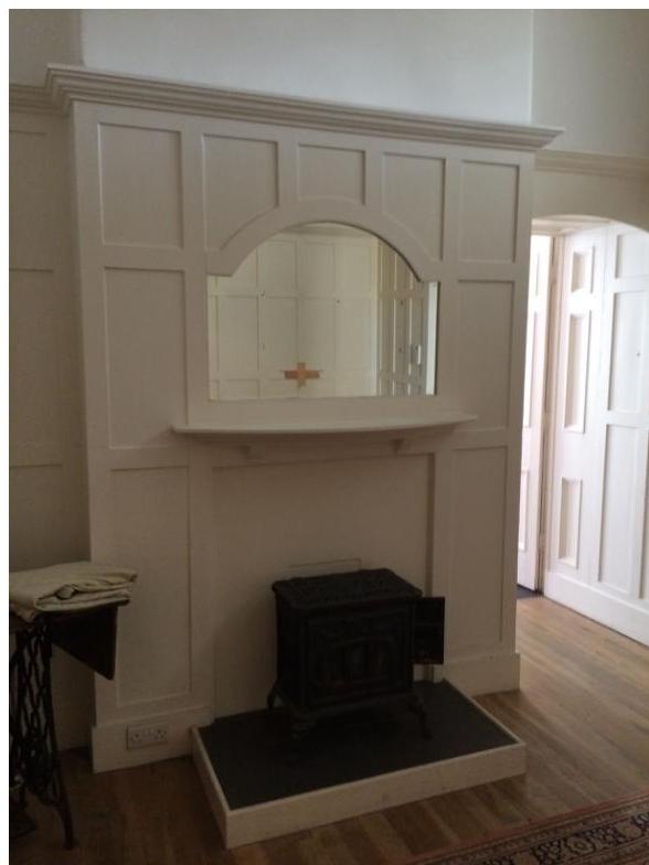 Interior photograph showing a white panelled fireplace surround with an inset mirror and a black stove on a hearth. An arched doorway to the right reveals white panelled doors in an adjacent room.