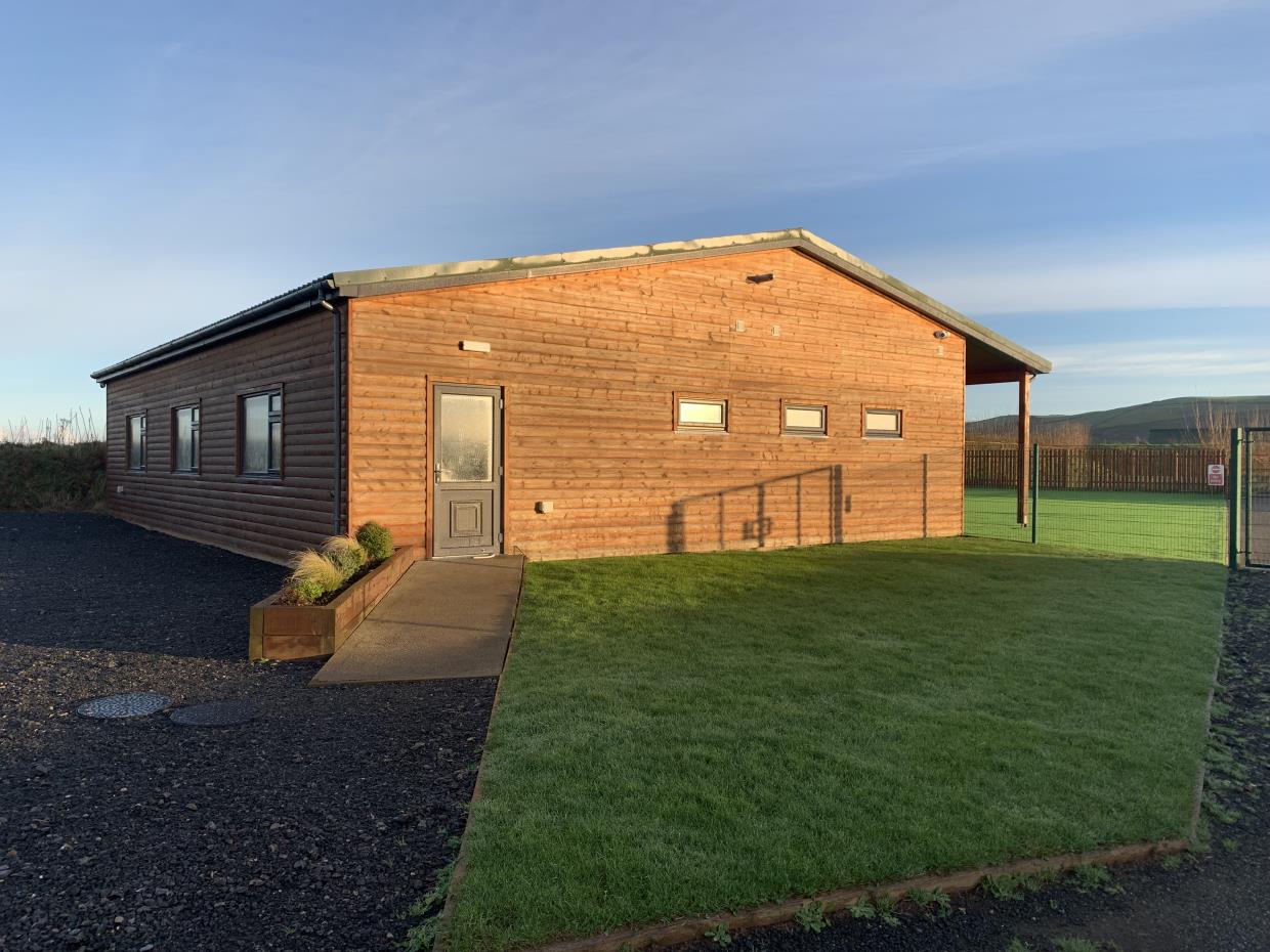 A photograph showing the exterior of a single-story wooden building with horizontal cladding and a grey door. The building is situated in a rural setting with a gravel driveway and a grassy area in the foreground.