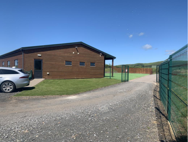 A photograph showing a single-story building with brown cladding, a gravel driveway, a parked car, and a green fence in a rural setting.
