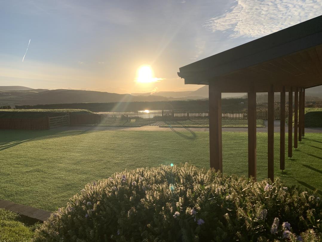 A photograph showing a wooden pergola structure alongside a manicured lawn with a scenic view of hills and water in the background. The image captures a rural setting with landscaping and a bright sun low on the horizon.