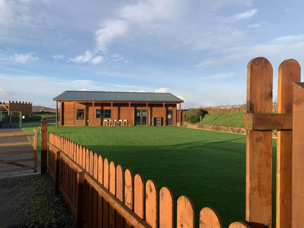A single-story wooden building with a dark roof stands behind a large green lawn, viewed through a wooden fence in the foreground.